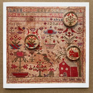 May include: A square sampler featuring cross-stitched alphabet letters, numbers, birds, flowers, and a house on aged fabric. Three circular glass cabochons with matching cross-stitch motifs are placed over the sampler, which reads Sarah Young Aged 11 Years.