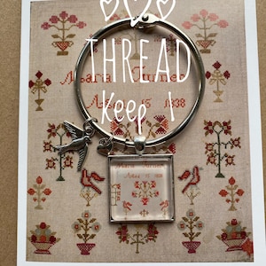 Puede incluir: Un porta hilos de plata con un pequeño encanto de pájaro y un encanto cuadrado de vidrio con el texto "Maria Turner, Aged 15, 1838" sobre un fondo blanco con un patrón floral rojo y verde.