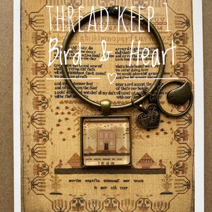 Puede incluir: Un sampler enmarcado con una casa y árboles bordados en tonos de rosa, marrón y verde. El sampler está titulado "martha augufta simmonds wer work in sev uth year". El sampler se muestra con un aro de metal, un amuleto de corazón y un amuleto de pájaro.