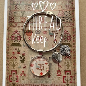 May include: A silver thread keeper with a round hoop, a heart charm, and a glass charm featuring a peacock design. The text "THREAD Keep" is written on the hoop. The background is a vintage fabric with a floral pattern.