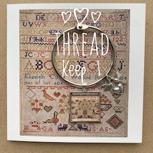 Op de afbeelding: Een zilveren draadhouder met een klein vierkant frame met een borduurwerkje. Het borduurwerkje toont een boom met vogels en een hart. De draadhouder heeft de tekst "THREAD Keep" erop.