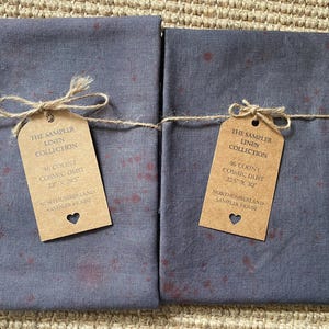 Pode incluir: Duas peças de tecido de linho cinzento-escuro dobradas com um padrão vermelho salpicado. Cada peça está atada com fio e apresenta uma etiqueta de papel castanho com a inscrição The Sampler Linen Collection, 46 Count Cosmic Dust, e Northumberland Sampler House.