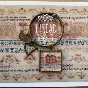 Puede incluir: Un amuleto de estilo vintage con un pequeño pájaro, un corazón y un amuleto cuadrado de vidrio con un diseño de casa roja y blanca. El amuleto está unido a un llavero con las palabras "THREAD Keep" escritas en él.