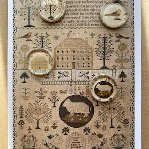 May include: A rectangular card featuring a beige sampler print with black and brown motifs of a house, trees, birds, and sheep. Four circular glass cabochons are placed on the card, displaying miniature images of a tree, a bird, a house, and a sheep.