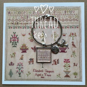 May include: A silver-tone bracelet with charms, including a dove, a heart, and a small framed cross-stitch design. The bracelet is displayed over a vintage cross-stitch sampler with the text "THREAD keep it".