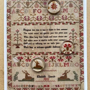 May include: A rectangular sampler featuring embroidered alphabets, floral motifs, and the text "Fragrant the rose is but it fades in time" alongside three circular glass cabochons displaying miniature cross-stitch patterns of a star, flower, and deer.