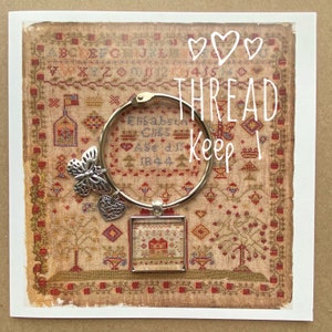 Könnte beinhalten: Ein silberner Fadenhalter mit einem Schmetterlingsanhänger, einem Herzanhänger und einem quadratischen Glasanhänger mit einem Vintage-Stickbild eines Hauses. Der Text "THREAD KEEP" ist in Weiß auf braunem Hintergrund geschrieben. Der Hintergrund ist ein Vintage-Stickmuster mit dem Text "Elisabeth Chess Age 17 1844".