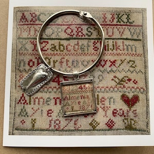 Puede incluir: Un anillo de plata con un dije de corazón y un pequeño marco cuadrado con el texto "45 Almenia 182" sobre un fondo beige con bordado de punto de cruz rojo y verde.