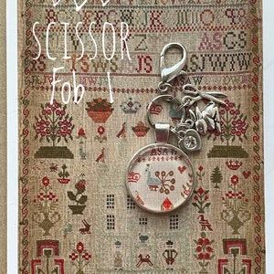 May include: A silver scissor fob with a charm of a rabbit and a bird. The fob is attached to a small round glass charm with a cross-stitch design. The fob is displayed on a vintage sampler with the text "SCISSOR Fob I" and "AMISOUTAR AGED 13 1832".