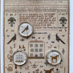 May include: A beige fabric sampler featuring cross-stitched text, birds, a house, and animals, overlaid with three circular silver-rimmed glass cabochons. The cabochons display a bird, a house, and a horse, mirroring motifs from the sampler background.