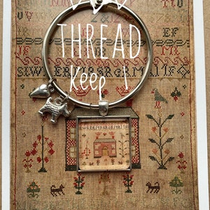 May include: A silver thread keeper with two hearts and a dog charm. The thread keeper is hanging from a small framed sampler with a house and the text "wreaths & berries".