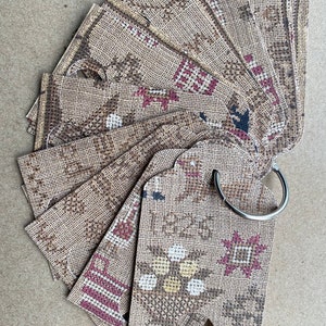 May include: A collection of cross-stitch design tags, featuring floral and geometric patterns in brown, white, and burgundy. The tags are held together by a silver metal ring. One tag displays the year "1825".