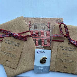 May include: A cross-stitch sampler kit featuring the text "ANN INNES c1830 HOUSE SNIPPIT" and "NORTHUMBERLAND SAMPLER HOUSE". The kit includes fabric, a pin, and a tag. The pin has a paisley design. The kit is tied with a red and blue ribbon.