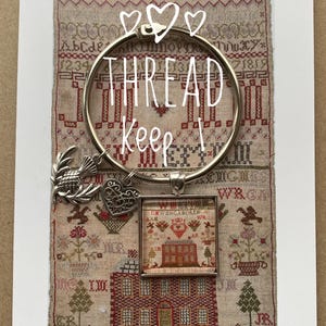 May include: A silver thread keeper with a heart charm and a small framed cross-stitch of a house. The text "THREAD KEEP" is written on the ring. The background is a vintage sampler with a floral pattern.
