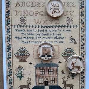 May include: A rectangular sampler featuring cross-stitch embroidery on beige fabric with an alphabet, a poem, and motifs of houses, deer, and flowers. Three circular silver-rimmed pins are placed on top, displaying a butterfly, a house, and a deer.
