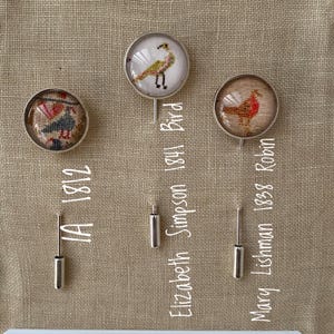May include: Three silver pin brooches with glass cabochons featuring embroidered bird designs. The brooches are labeled with the maker's name and date: "Mary Lishman 1838 Robin", "Elizabeth Simpson 1841 Bird", and "HA 1812".