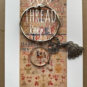 May include: A white card featuring a printed image of an antique sampler with a colourful floral design. The card has a silver ring with the words "THREAD Keep" written on it. The ring is attached to a silver charm with a peacock design and a small glass charm with a colourful bird design.