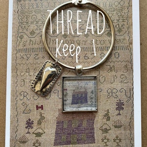 Puede incluir: Un porta hilos de plata con un dije de corazón y una pequeña pieza de punto de cruz enmarcada con el texto "1872" y las palabras "THREAD Keep".