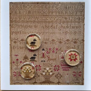 May include: A square piece of tan linen fabric featuring a cross-stitch alphabet, numbers, and decorative motifs including a person, animals, and flowers. Three circular silver-rimmed brooches with cross-stitch inserts are placed on the fabric.