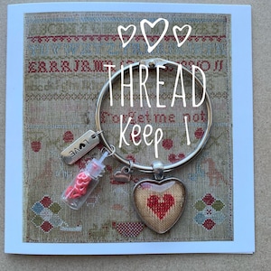 May include: A silver keyring with the words "THREAD Keep" in white script. It features a small vial of red thread, a heart charm, and a heart-shaped pendant with a stitched red heart design. The background is a cross-stitch pattern.