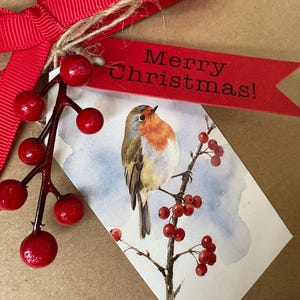 May include: A gift tag featuring a watercolor illustration of a robin perched on a berry branch, accented with a red ribbon, a sprig of artificial red berries, and a red paper tag with the black printed text Merry Christmas! on brown paper.