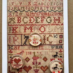 May include: A rectangular sampler featuring rows of embroidered letters, numbers, and motifs in red, black, and brown thread on beige fabric. Three circular glass cabochons are placed on the sampler, displaying magnified details of the embroidery.