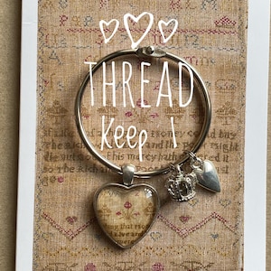 Op de afbeelding: Een zilveren draadring met een hartvormige bedel, een kroonbedel en een hartbedel. Op de ring staat de tekst "THREAD Keep!". De achtergrond is een beige stof met een bloemenpatroon.