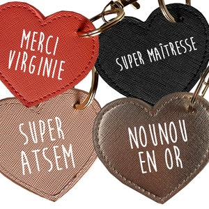 May include: Four heart-shaped keychains with different messages. The first keychain is red and says "Merci Virginie". The second keychain is black and says "Super Maitresse". The third keychain is pink and says "Super Atsem". The fourth keychain is gold and says "Nounou en Or".