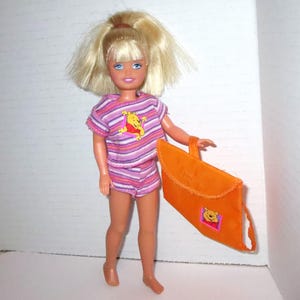 May include: A doll with blonde hair wearing a pink and purple striped shirt and shorts. The shirt features a Winnie the Pooh graphic. The doll is holding an orange bag with a Winnie the Pooh patch.