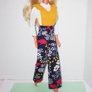 May include: A doll wearing a white long-sleeved shirt, a mustard yellow knit vest, and wide-leg pants. The pants are navy blue with a floral pattern in white, red, and yellow. The doll has blonde hair.
