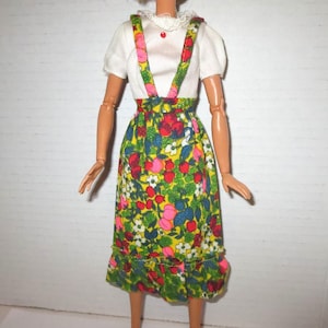 May include: A vintage doll wearing a white blouse with a lace collar and a red button. The doll is also wearing a floral print skirt with suspenders. The skirt features a yellow background with red, pink, and blue flowers.