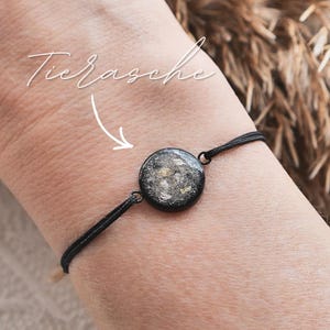 Könnte beinhalten: Schwarzes Kordelarmband mit einem runden, dunklen Anhänger mit hellen Flecken. Der Anhänger ist der Mittelpunkt und ist mit kleinen Metallschlaufen mit dem Kordel verbunden. Das Armband wird am Handgelenk getragen.
