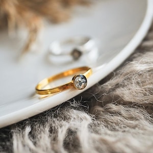 Könnte beinhalten: Ein goldener Ring mit einem kleinen, runden, mehrfarbigen Stein in einer Lünettenfassung. Der Ring liegt auf einer weißen Oberfläche, mit einem zweiten Ring unscharf im Hintergrund. Der Hintergrund hat eine weiche, neutrale Farbe.