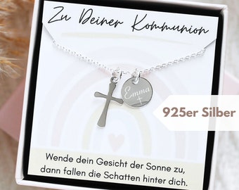 Może przedstawiać: Srebrny naszyjnik z krzyżykiem i okrągłym wisiorkiem z wygrawerowanym imieniem "Emma" i krzyżem. Naszyjnik jest prezentowany w białym pudełku z napisem "Zu Deiner Kommunion" i "925er Silber". Tło zdobi tęczowy wzór.