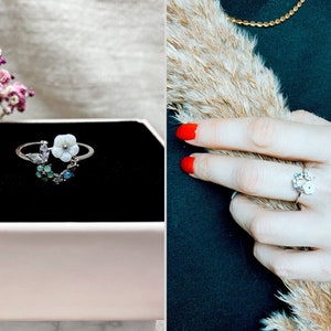 Op de afbeelding: Een zilveren ring met een witte bloem en groene en blauwe edelstenen. De ring ligt op een zwarte achtergrond.