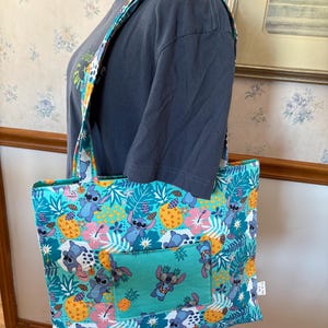May include: A teal tote bag with a colorful pattern featuring cartoon characters, pineapples, and tropical flowers. The bag has a matching strap and a front pocket with a smaller version of the design. The bag is on a mannequin.