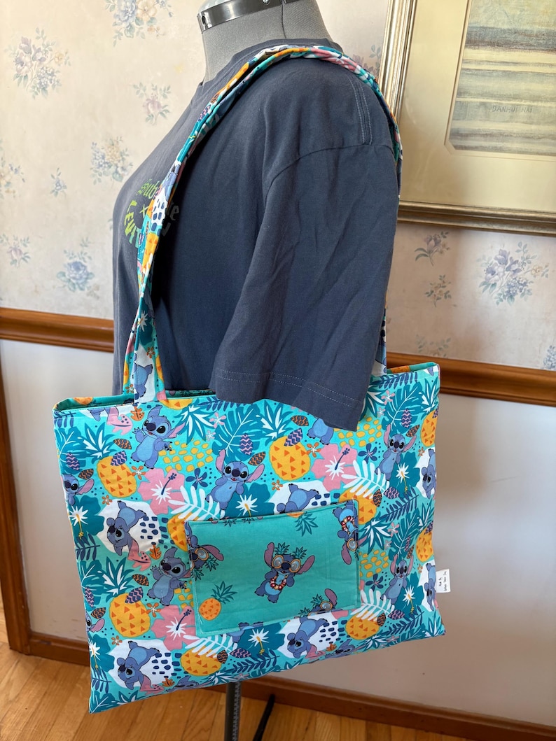 May include: A teal tote bag with a colorful pattern featuring cartoon characters, pineapples, and tropical flowers. The bag has a matching strap and a front pocket with a smaller version of the design. The bag is on a mannequin.