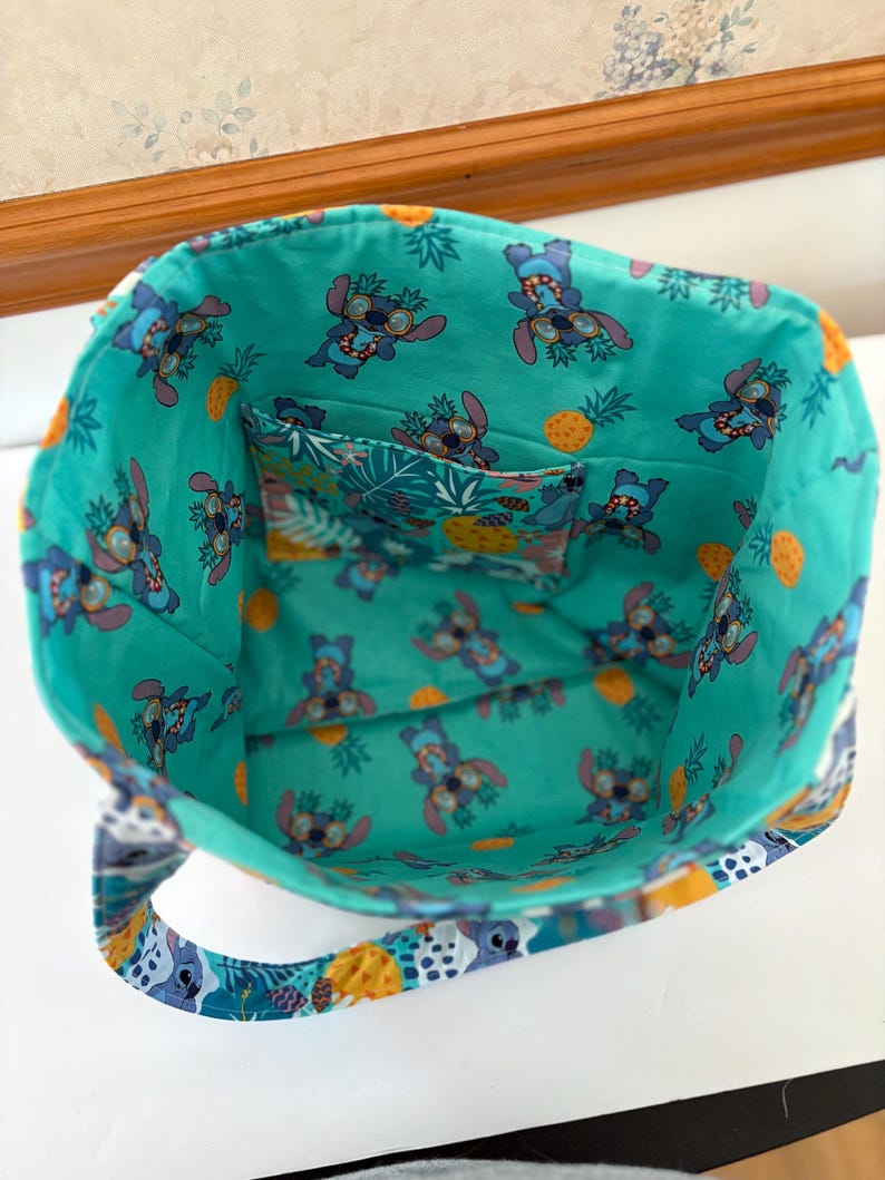 May include: A turquoise tote bag with a colorful interior featuring a cartoon character wearing sunglasses and pineapples. The bag has a matching interior pocket and handles with a patterned design. The bag is open, showing the inside.