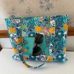 May include: A teal tote bag with a colorful pattern featuring cartoon characters, pineapples, and tropical flowers. The bag has a front pocket and handles. Sunglasses are placed in the front pocket.
