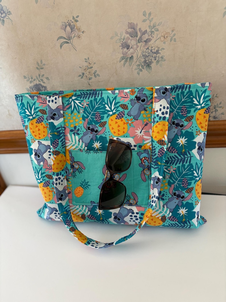 May include: A teal tote bag with a colorful pattern featuring cartoon characters, pineapples, and tropical flowers. The bag has a front pocket and handles. Sunglasses are placed in the front pocket.