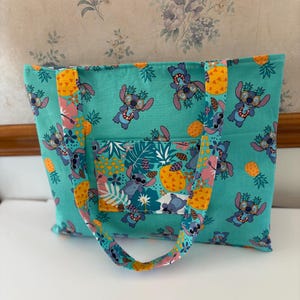 May include: A teal tote bag featuring a repeating pattern of a cartoon character wearing sunglasses and leis, pineapples, and tropical leaves. The bag has a front pocket and matching handles.
