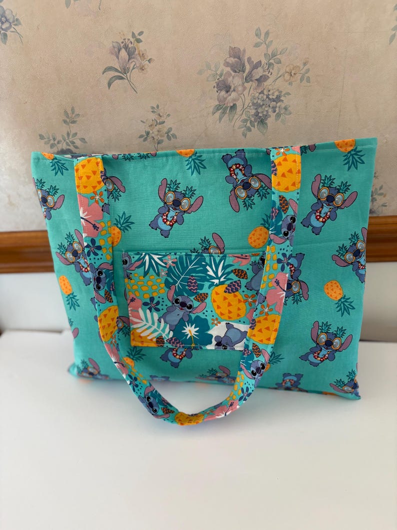 May include: A teal tote bag featuring a repeating pattern of a cartoon character wearing sunglasses and leis, pineapples, and tropical leaves. The bag has a front pocket and matching handles.