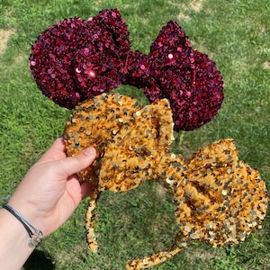 May include: A pair of sequined Minnie Mouse ears with a burgundy bow and a gold bow.