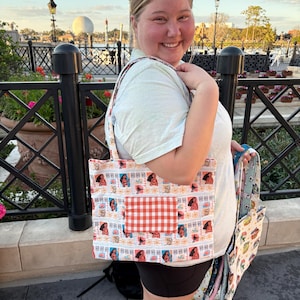 Puede incluir: Un bolso tote estampado con un dise&ntilde;o repetido de una mujer en un sello postal. El bolso tiene un bolsillo a cuadros rojos y blancos. El bolso lo sostiene una persona que lleva una camiseta azul claro y pantalones cortos negros.