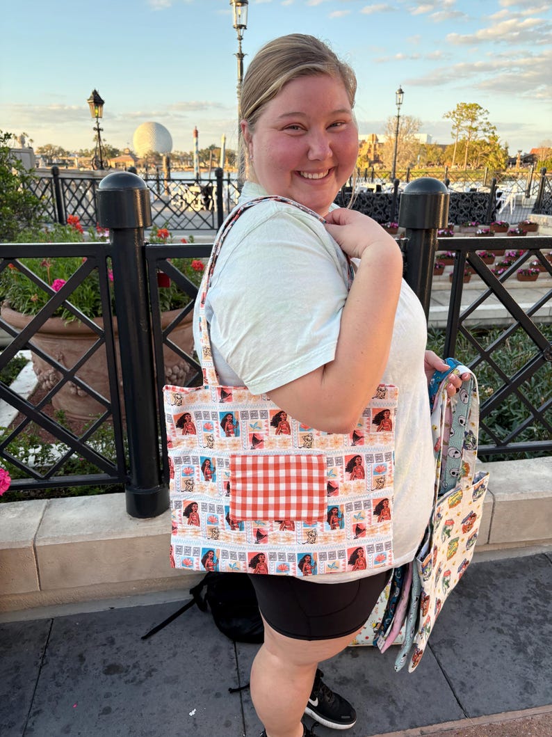 Puede incluir: Un bolso tote estampado con un dise&ntilde;o repetido de una mujer en un sello postal. El bolso tiene un bolsillo a cuadros rojos y blancos. El bolso lo sostiene una persona que lleva una camiseta azul claro y pantalones cortos negros.