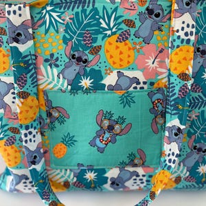 May include: A teal tote bag featuring a repeating pattern of the character Stitch, pineapples, and tropical flowers. The bag has a teal pocket with Stitch wearing sunglasses. The handles and trim are yellow.