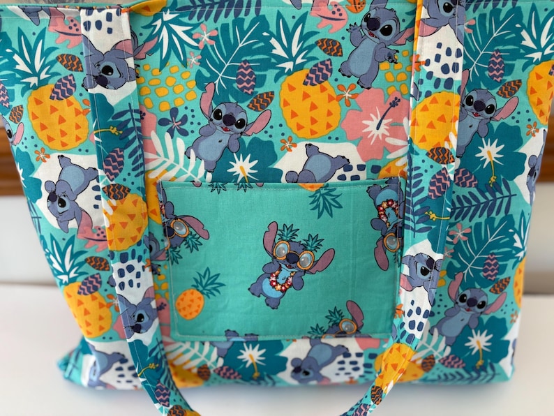 May include: A teal tote bag featuring a repeating pattern of the character Stitch, pineapples, and tropical flowers. The bag has a teal pocket with Stitch wearing sunglasses. The handles and trim are yellow.