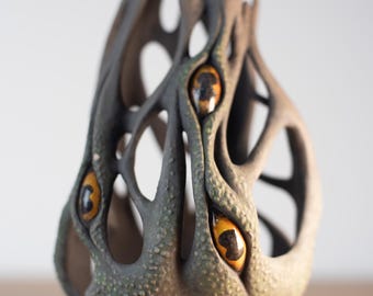 Könnte beinhalten: Eine Keramikvase mit einem strukturierten, baumartigen Design und drei gelben Augen. Die Vase ist dunkelgrün und braun.