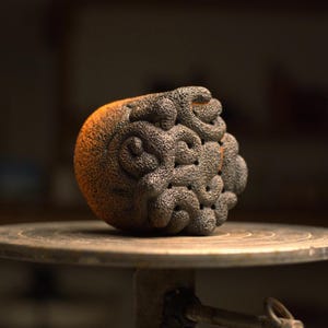 May include: A textured, spherical sculpture with a dark gray and orange color scheme. The surface features an intricate, swirling pattern. The sculpture is displayed on a round, wooden surface, suggesting a pottery or art piece.