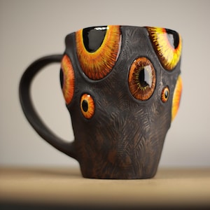 Puede incluir: Una taza de cerámica marrón oscuro con una superficie texturizada y un asa. La taza está adornada con múltiples diseños de ojos detallados en tonos naranja, amarillo y marrón. La taza está sobre una superficie de madera.